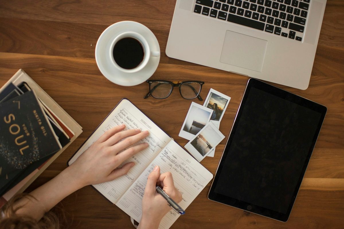 Persona escribiendo en un cuaderno junto a un café y un portátil, vista desde arriba