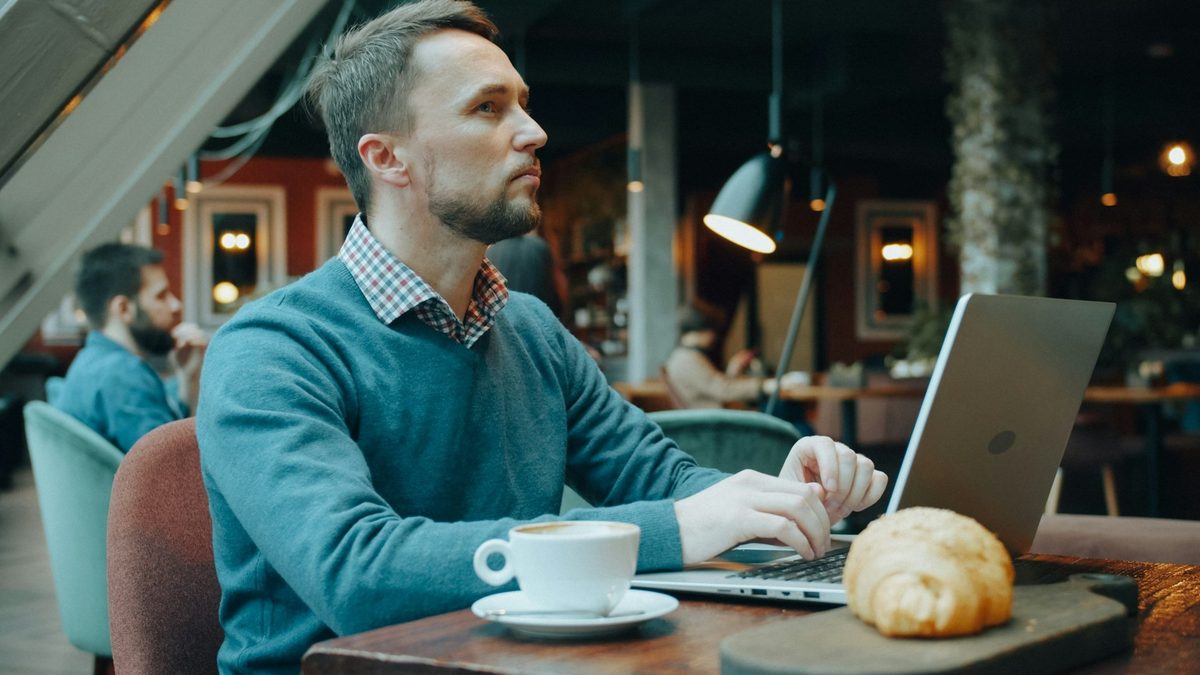 Hombre trabajando con un portátil en una cafetería, con un café y un cruasán