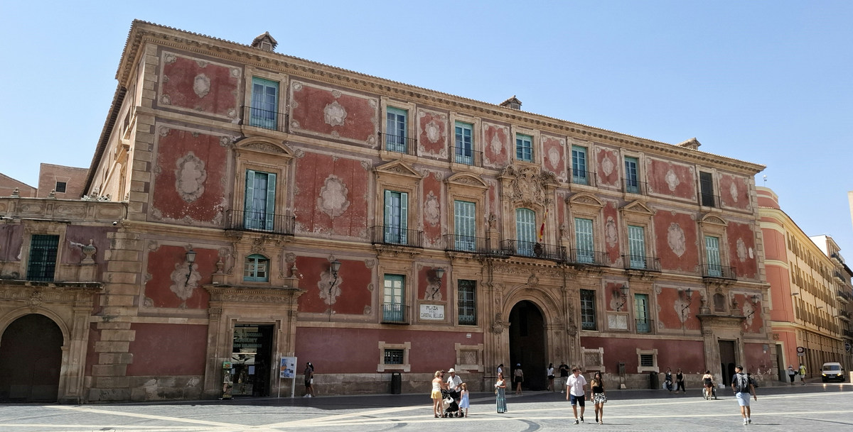 La fachada rojiza del Palacio Episcopal de Murcia, en la plaza del Cardenal Belluga, con gente paseando delante